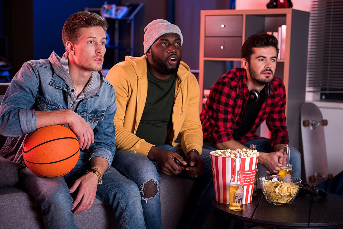 So exciting. Three young interested guys are sitting on sofa and expressing exhilaration while watching sport game with opened mouthes. Friends are eating popcorn and drinking beer