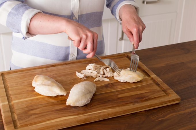 Person using two forks to shred chicken breasts on a wooden cutting board