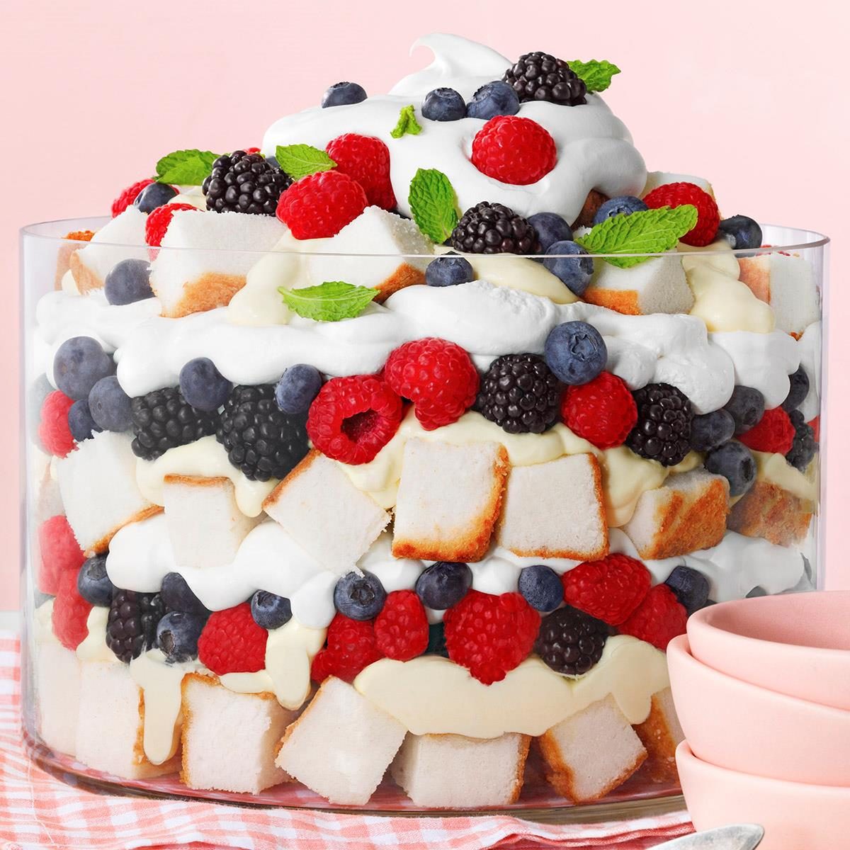 A layered trifle in a clear glass dish with angel food cake, whipped cream, blackberries, blueberries, and raspberries. Mint leaves garnish the top. Pink bowls are in the foreground.