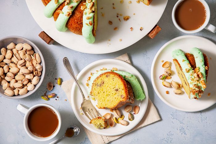 Piece of Pistachio Pudding Cake On A White Plate with A Bowl of Pistachio and Coffee Cups Next to it