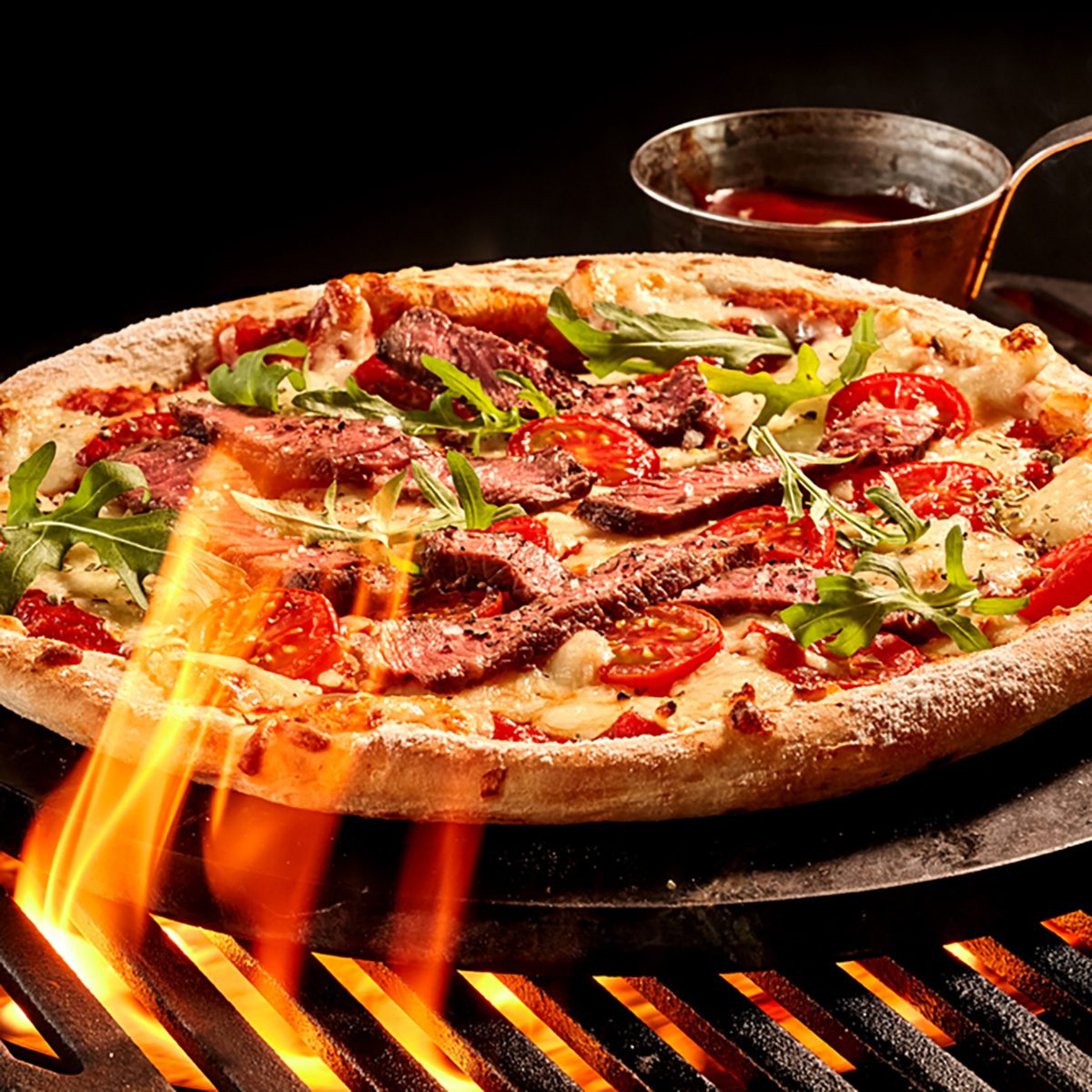 Flames reaching up toward meat and vegetable cheese pizza on grill. Hot sauce in bowl on iron plate