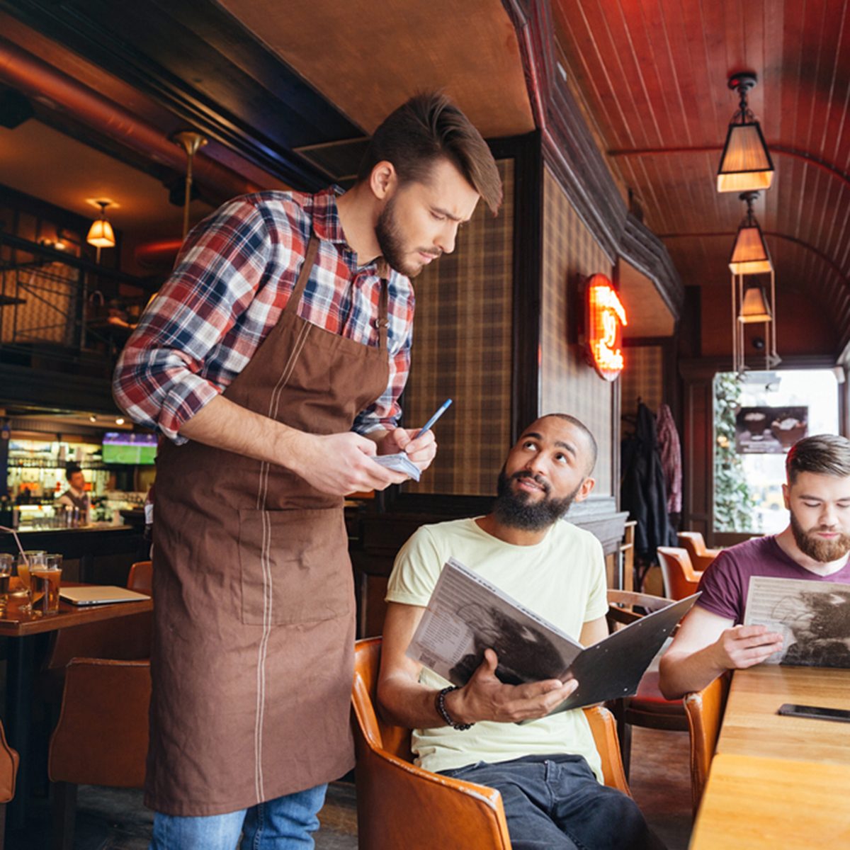 Serious concentrated young waiter standing and taking an order from two bearded handsome men in cafe
