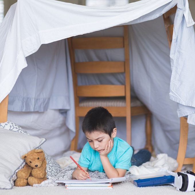 Young Native American boy draws in his sketchpad underneath his blanket fort in the living room