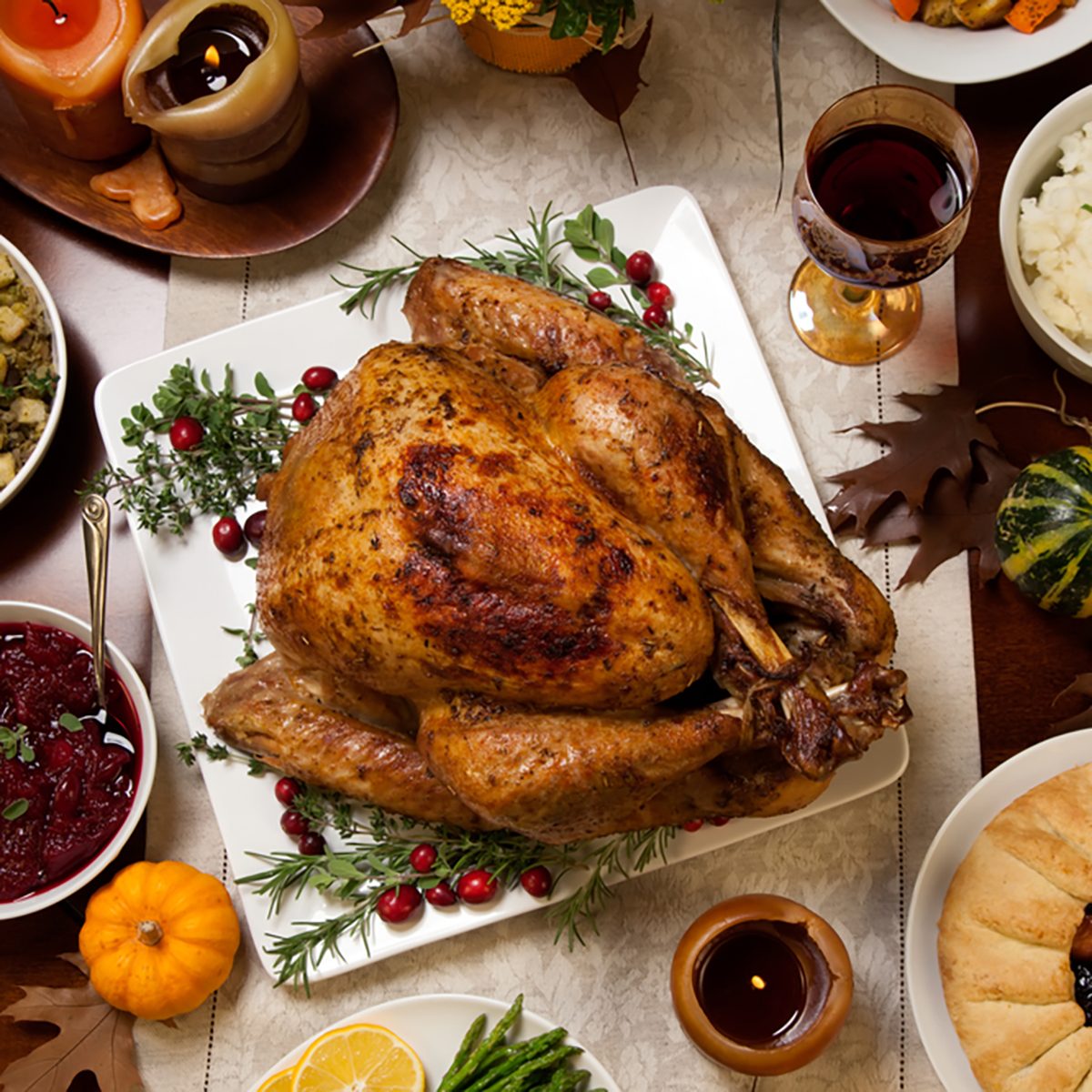 Roasted turkey garnished with cranberries on a rustic style table decoraded with pumpkins, gourds, asparagus, brussel sprouts, baked vegetables, pie, flowers, and candles.