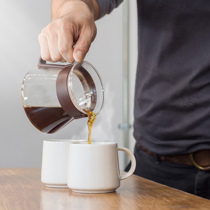 The man is pouring hot coffee from the coffee pot into the white coffee on the wooden table in the morning