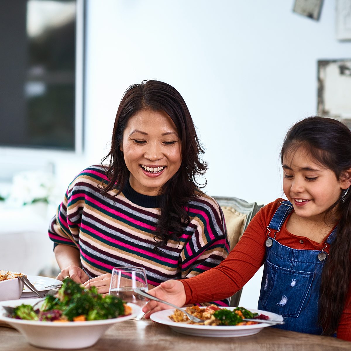 Cheerful family dinner in dining room, mid adult woman smiling at daughter eating food, good choice, positive parenting