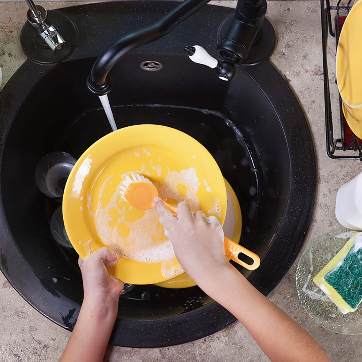 Child hands washing a plate in the kitchen sink - scrubbing with a brush under the water jet - top view