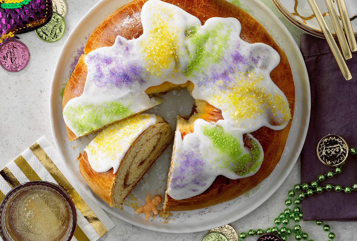 Traditional New Orleans King Cake; Overhead camer angle; White russian speckled surface; champagne; gold accents; gold flatware; Mardi Gras decorations; baby figure