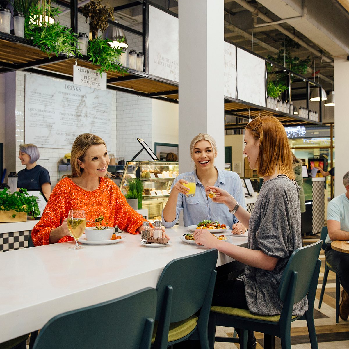 Three women are sitting at a table in a health cafe. They are eating organic, vegan meals while talking and laughing.
