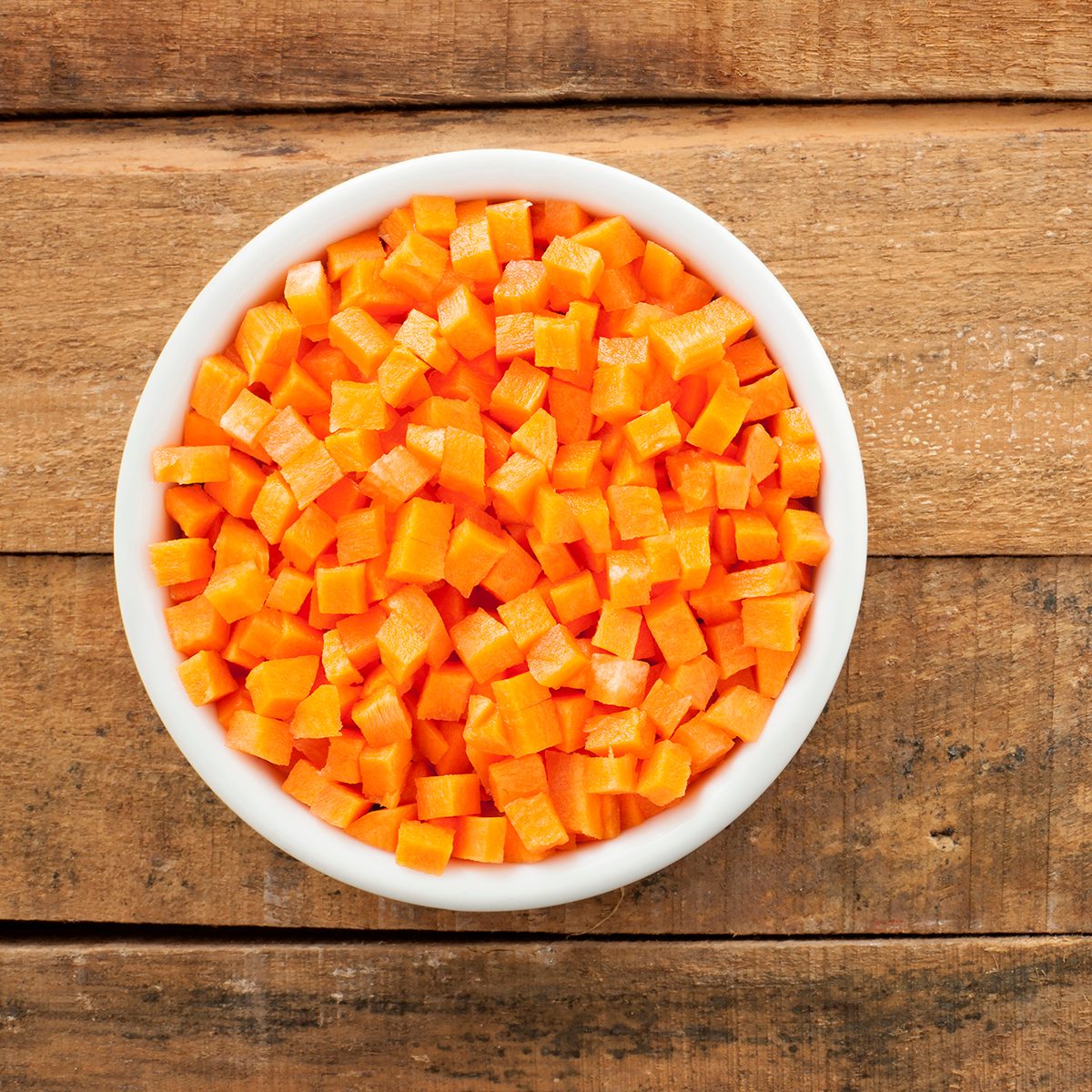Top view of white bowl full of small carrot cubes over wooden table