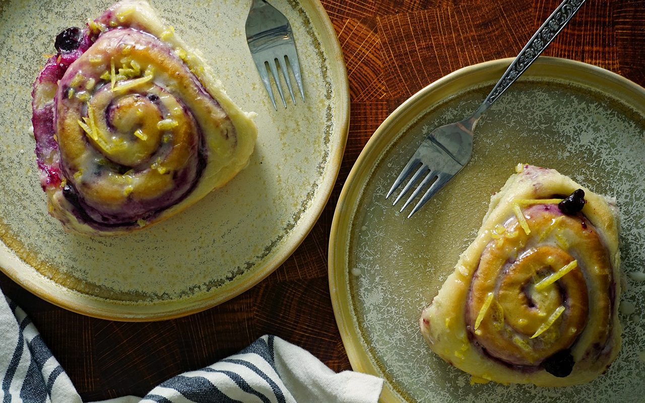 two baked blueberry sweet rolls from Magnolia Table, Vol. 2 on plates with lemon glaze and forks