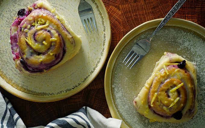 two baked blueberry sweet rolls from Magnolia Table, Vol. 2 on plates with lemon glaze and forks