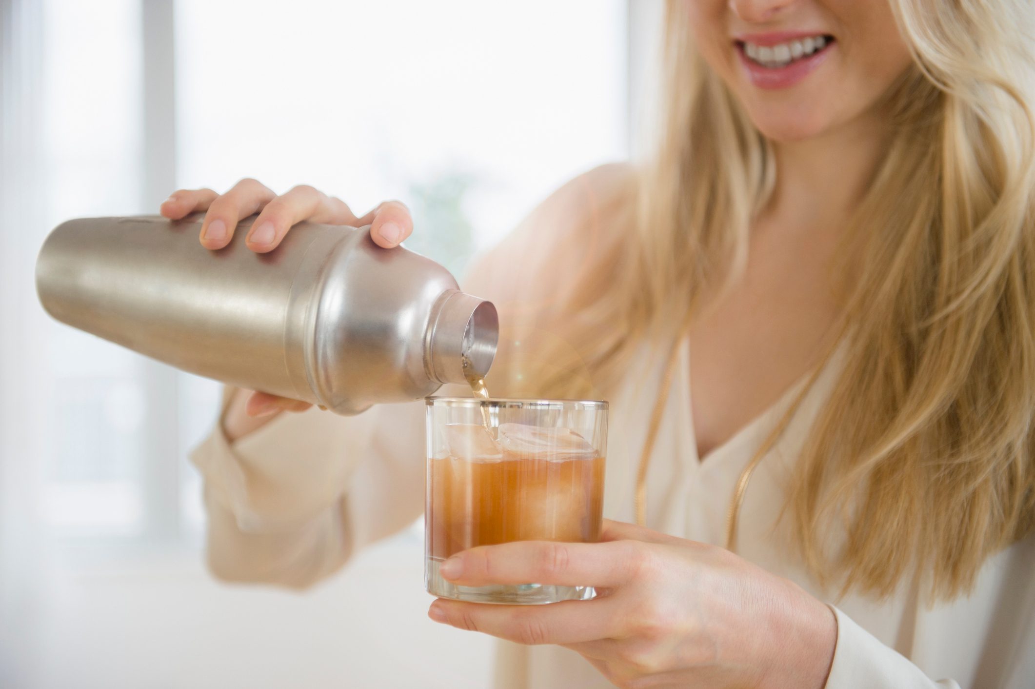 young woman pouring cocktail from cocktail shaker into glass at home