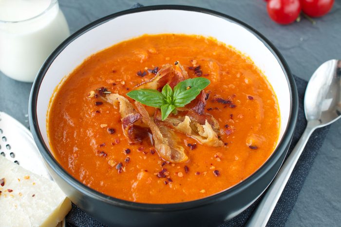 Tomato Soup With Basil And Fried Bacon In A Black Ceramic Bowl On A Grey Abstract Background