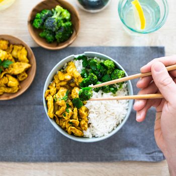Hand holding chopsticks with a bowl of rice, broccoli and chicken on a placemat