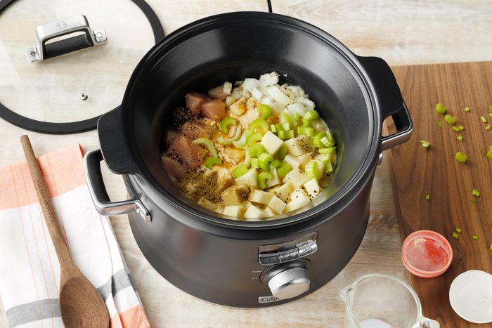 An electric slow cooker on a wooden surface with chopped vegetables, including onions, celery, and potatoes, as well as pieces of raw chicken inside. Nearby are a wooden spoon, an orange and white kitchen towel, and various small containers with ingredients.