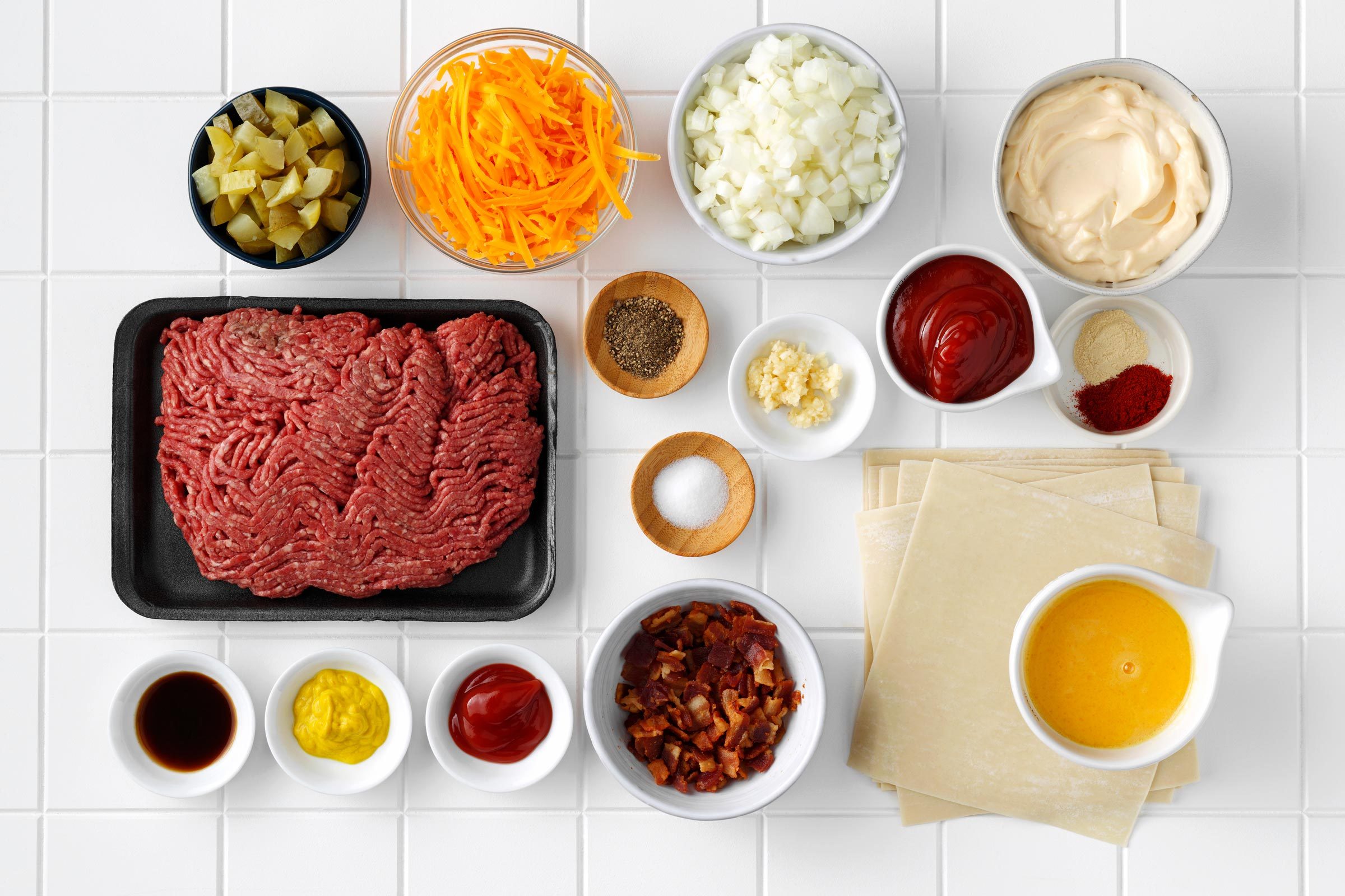 ingredients for cheeseburger egg rolls measured out into small bowls on a white tile kitchen counter