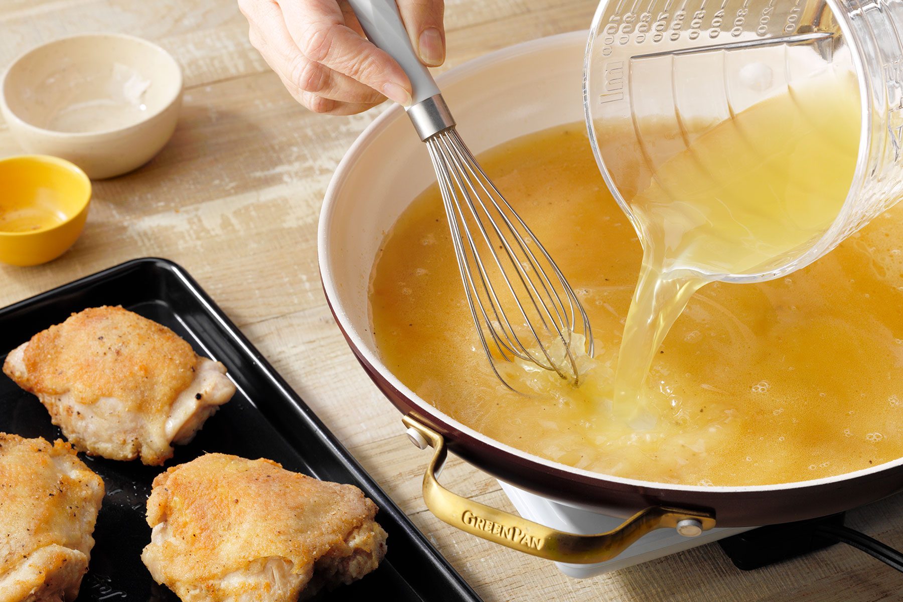 A person pours a clear yellow liquid from a measuring cup into a large saucepan on a stove, while whisking with a metal whisk. To the side, there is a baking tray with three cooked pieces of crispy brown chicken and small bowls with ingredients.