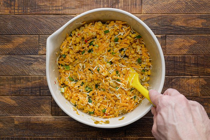 a person mixing canned corn, diced green onions, green chilies and diced jalapeno in bowl