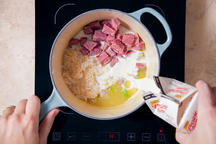 pouring milk into the saucepan filled with sauerkraut, corned beef and broth on Induction Cooktop on A Marble Kitchen Top