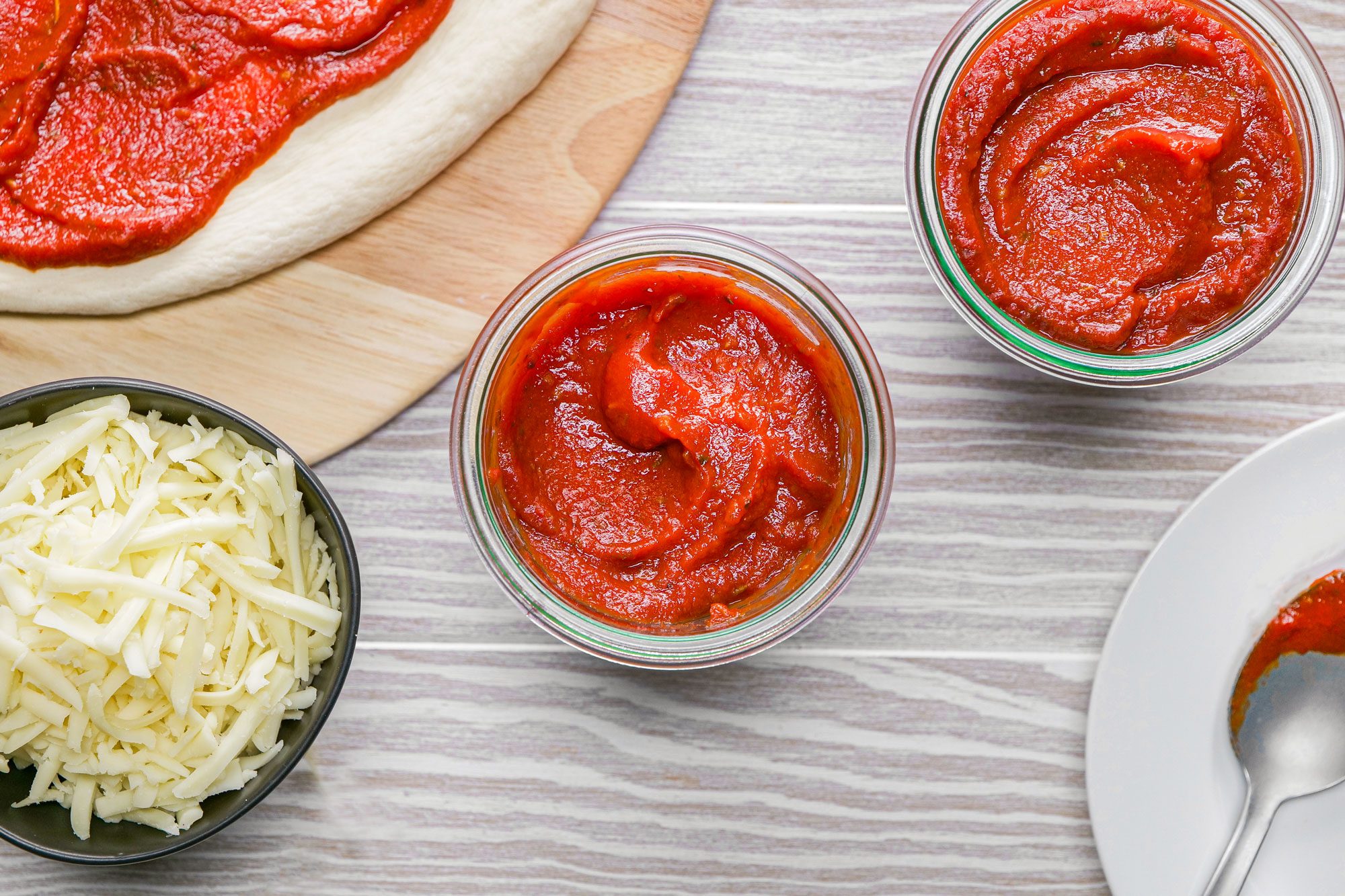 on a wooden surface Homemade pizza sauce in round glass containers and a bowl of cheese next to it