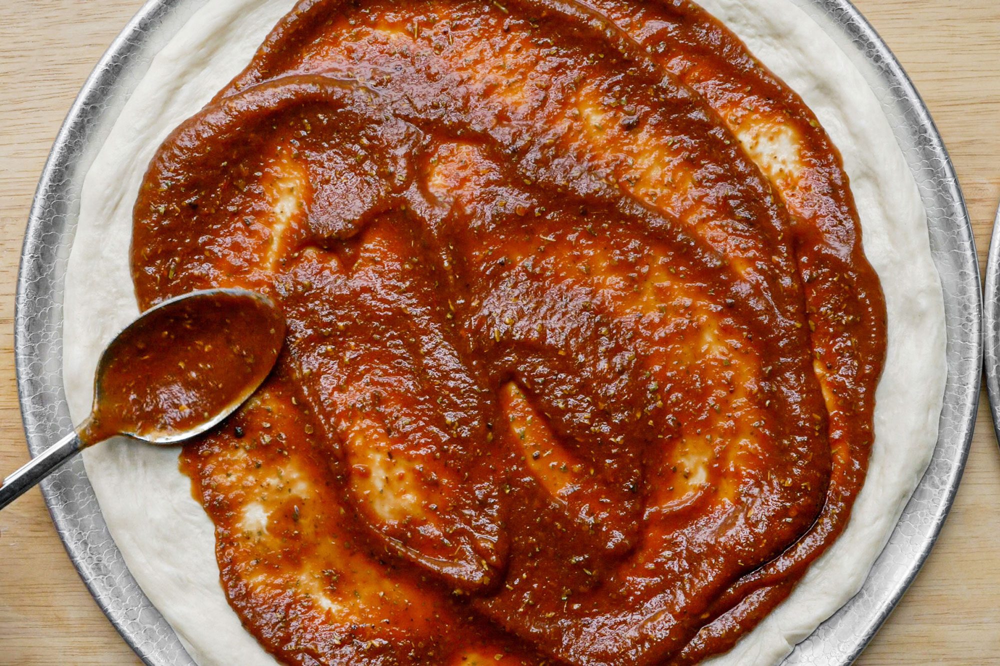 applying pizza sauce with a spoon on the pizza base placed on a plate.