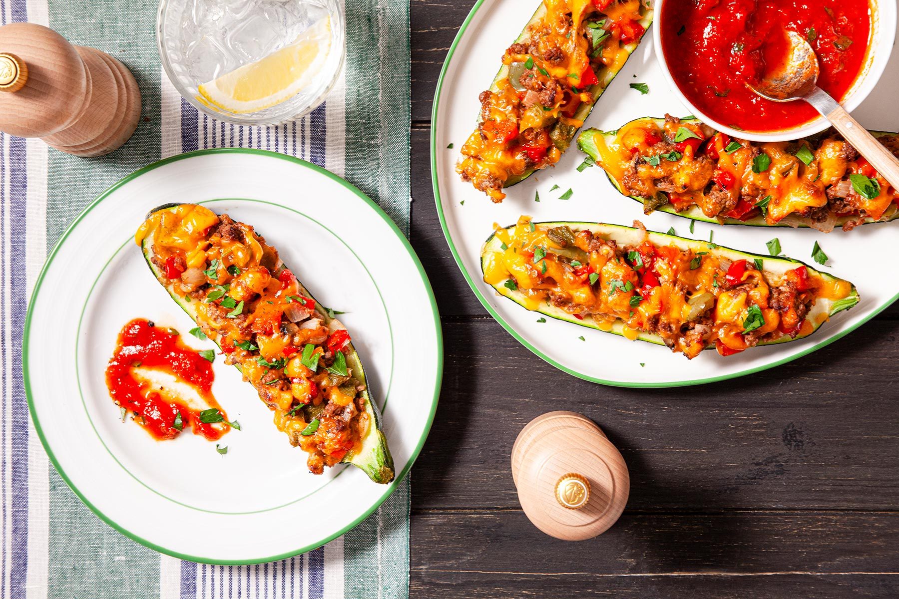 Overhead shot of Zucchini Boats served with marinara sauce and drink; salt shaker; spoon; bowl; table cloth; wooden background;