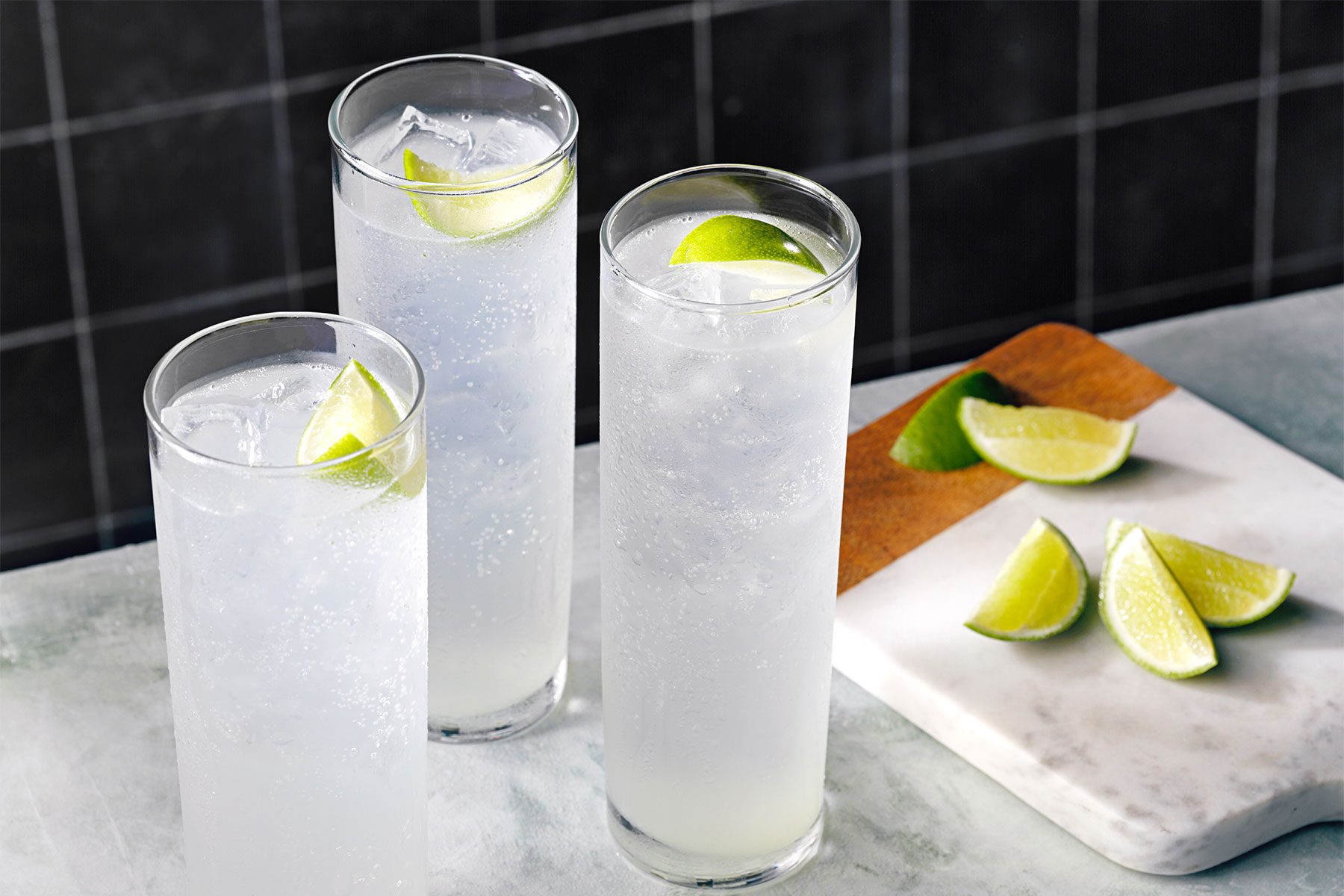 High angle shot of Gin Rickey; served in three collins glasses; garnished with lime wedge; lime wedges on chopping board; marble background;