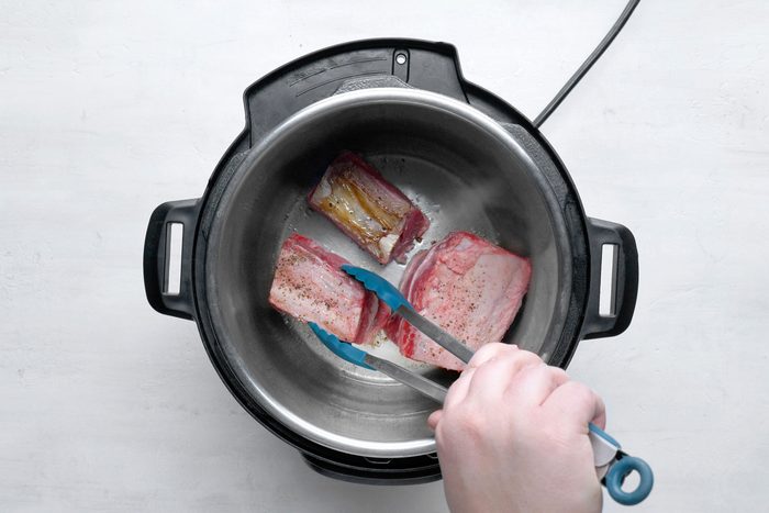 overhead shot; white background; cooking beef short ribs into electric pressure cooker using tong;
