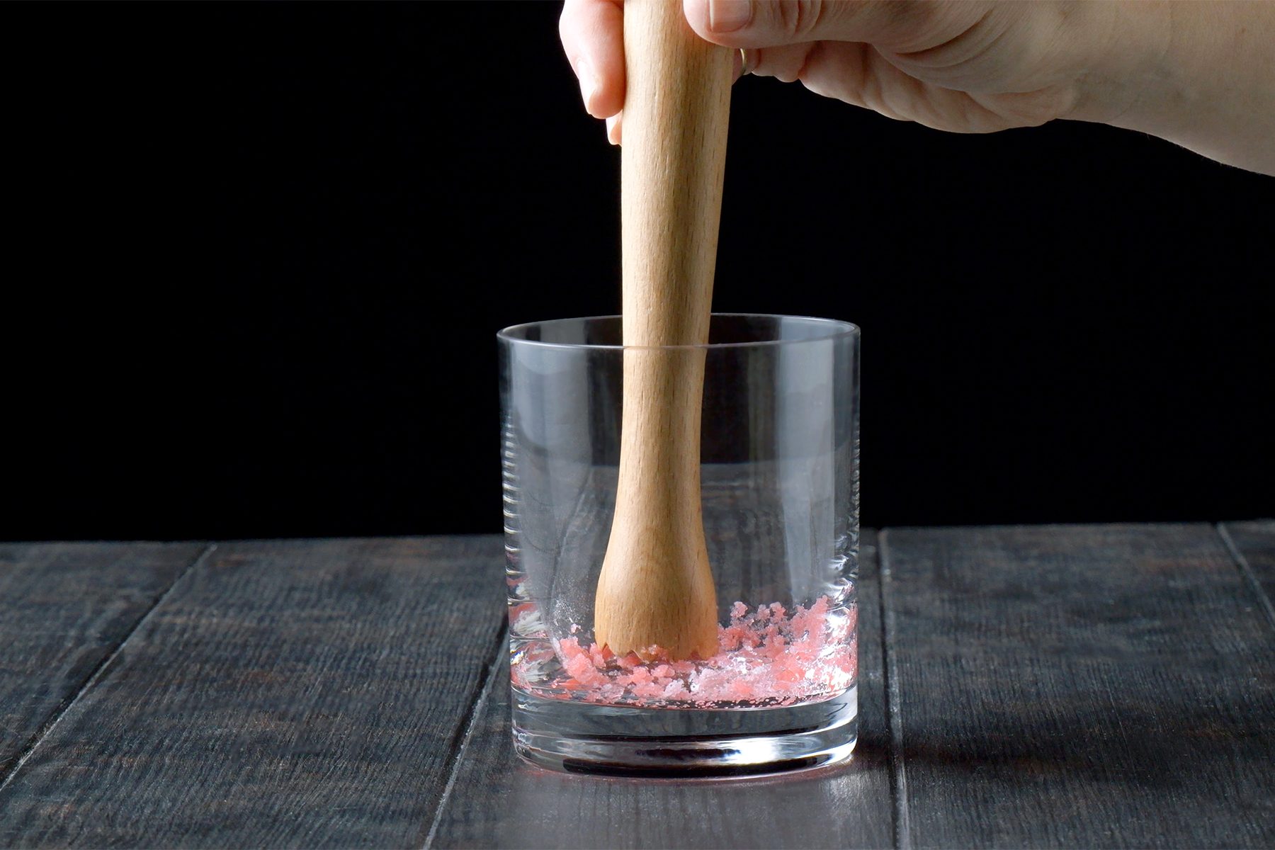 A hand is using a wooden muddler to crush pink salt crystals at the bottom of a clear glass.