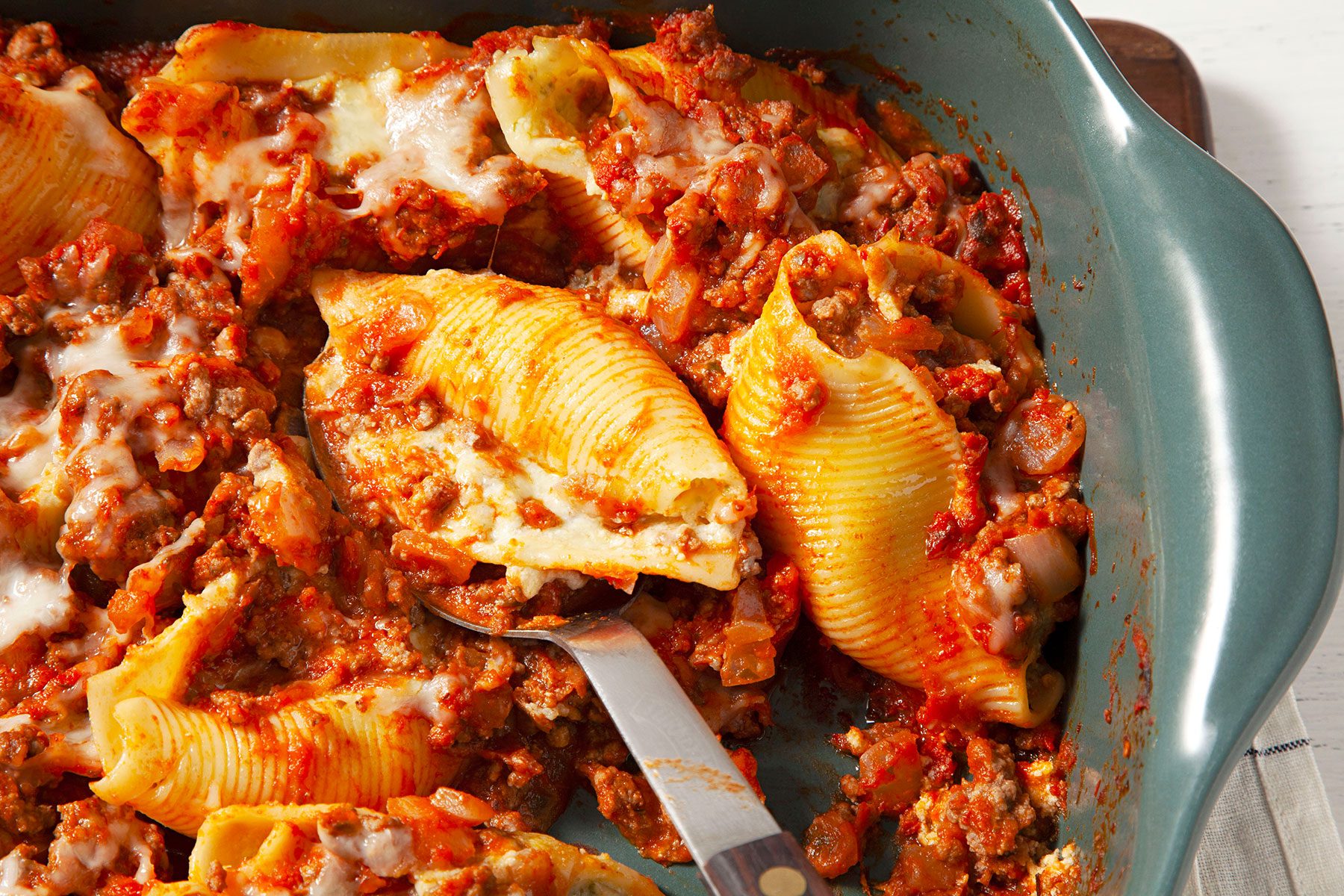 Close shot of Easy Beef-Stuffed Shells; baking dish; serving spoon; wooden tray; napkin; white wooden background;
