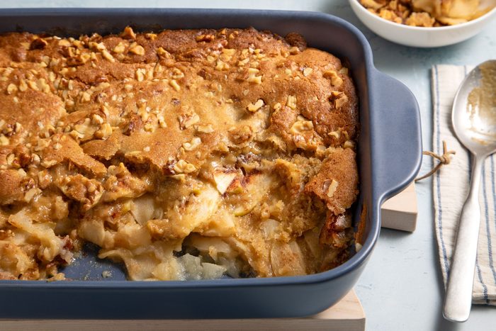 3/4th close shot; light blue textured background; Walnut Apple Dessert in a baking dish with silver spoon over kitchen towel;