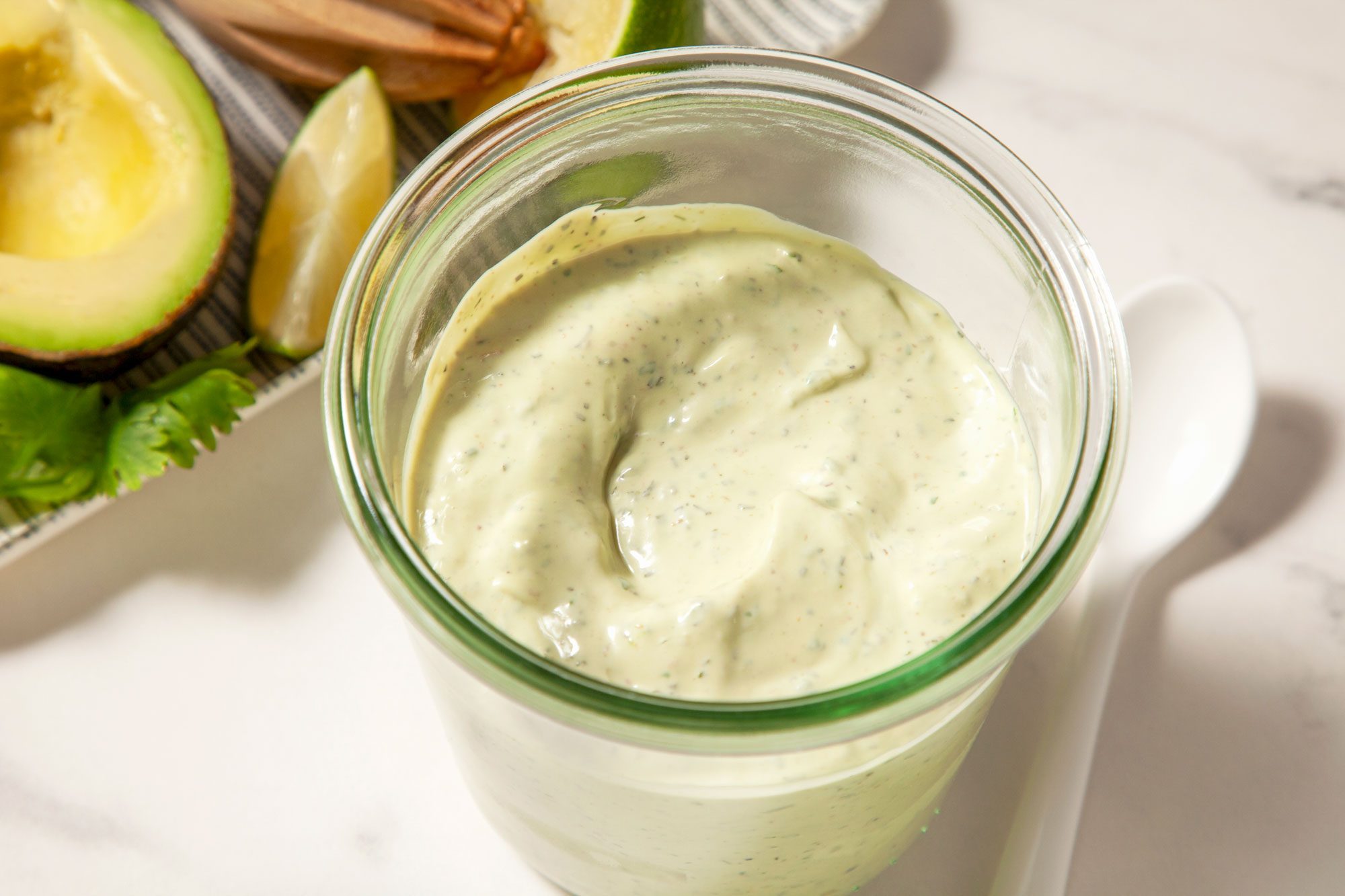 Top view hot of Avocado-Lime Ranch Dressing; in a glass jar; spoon; half cut avocado and lime wedges in a tray; marble background;