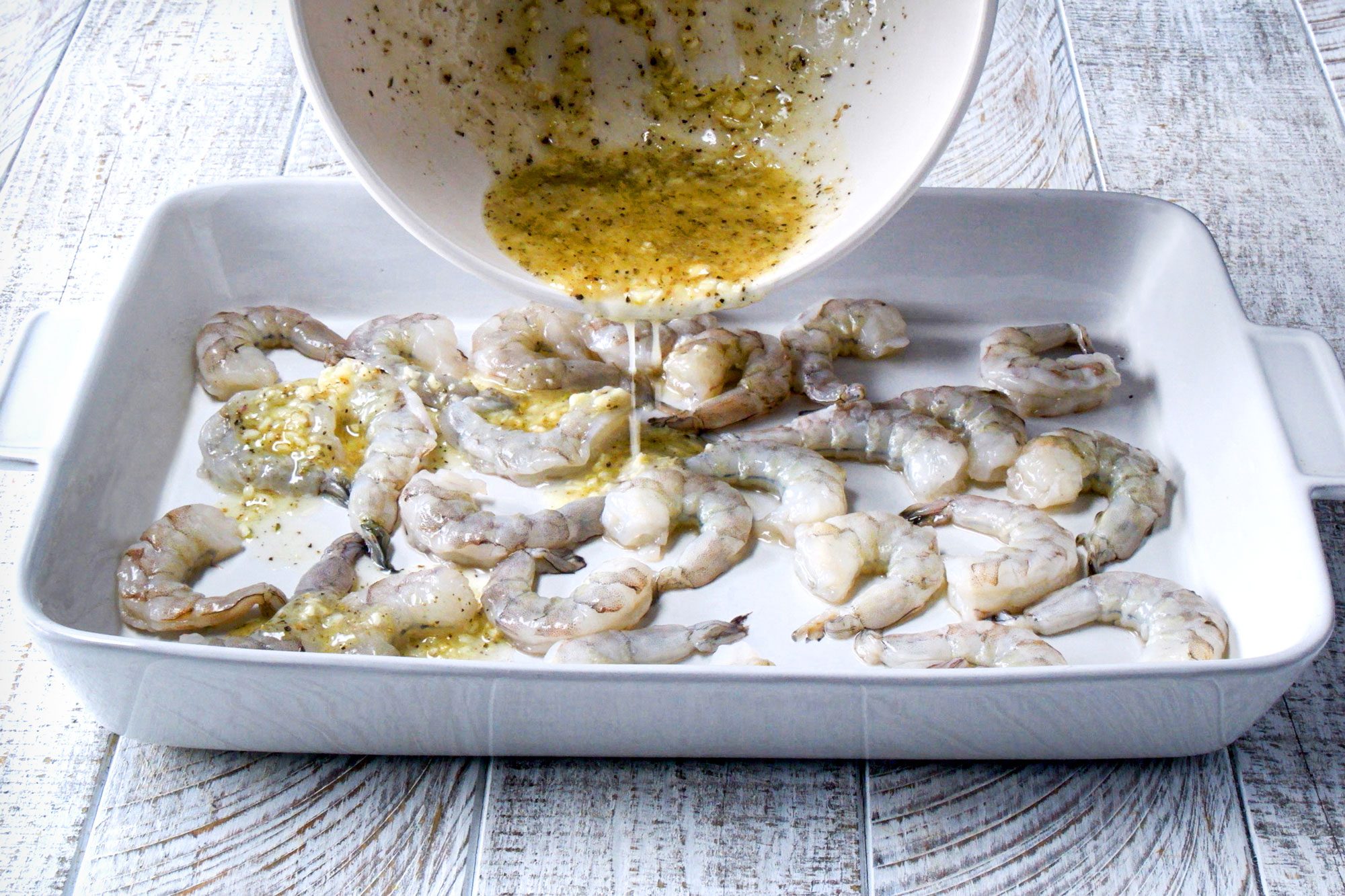 3/4th shot; white wooden background; pouring egg mixture over shrimps in rectangular dish; from white bowl;