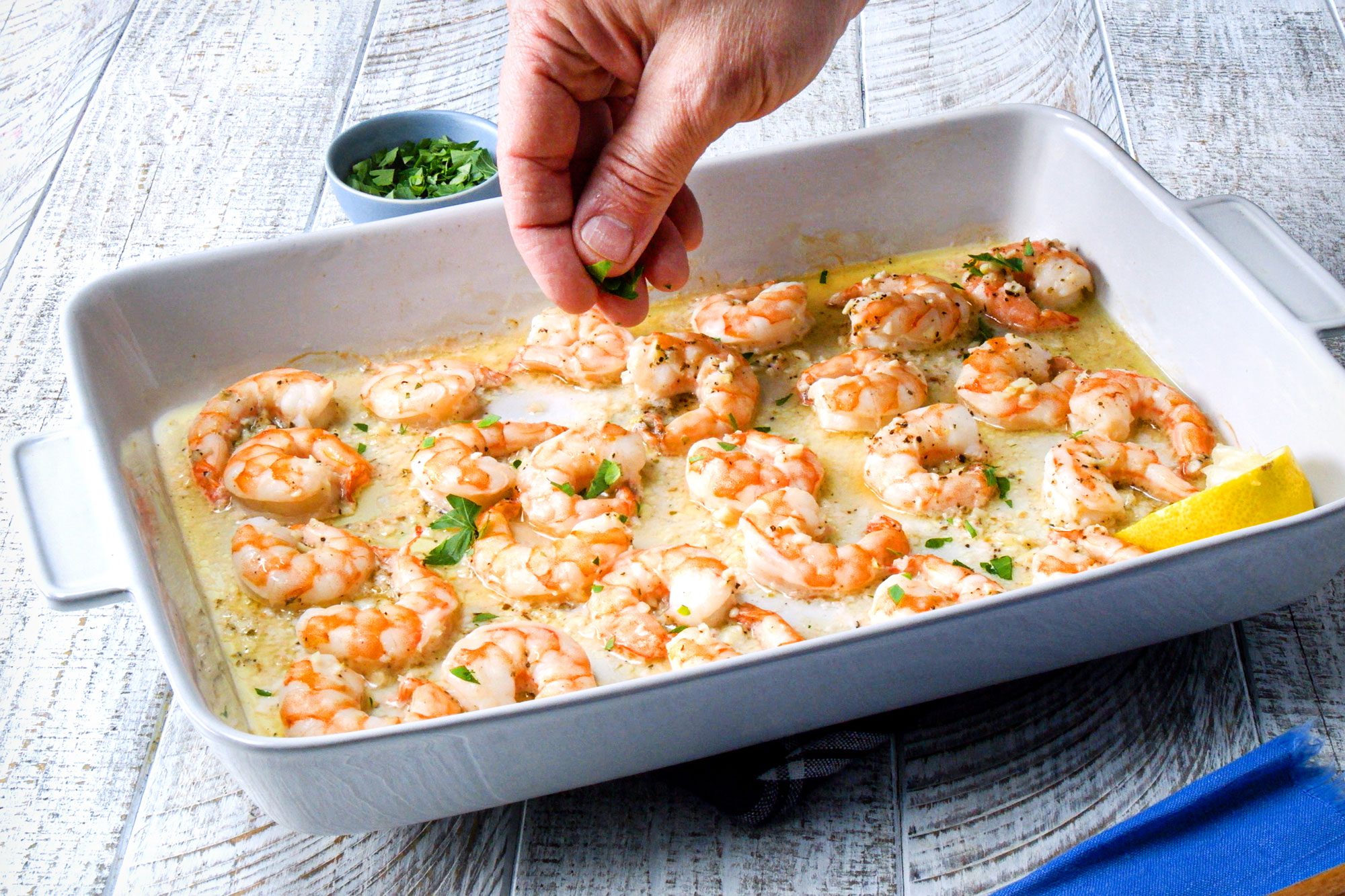 overhead shot; white wooden background; sprinkling chopped parsley over baked shrimps; in white rectangular dish;