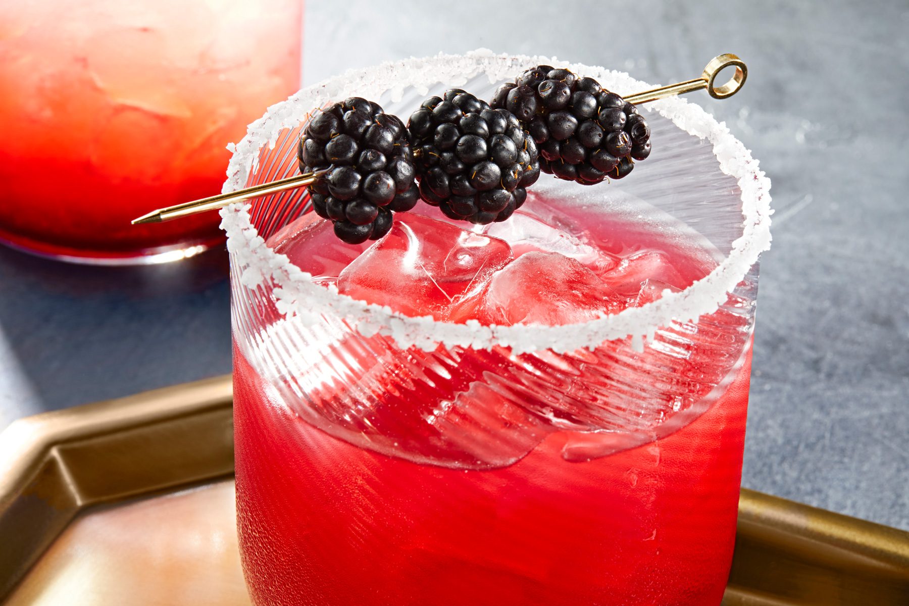 A blackberry margarita with a salted rim, garnished with blackberries on a golden tray.