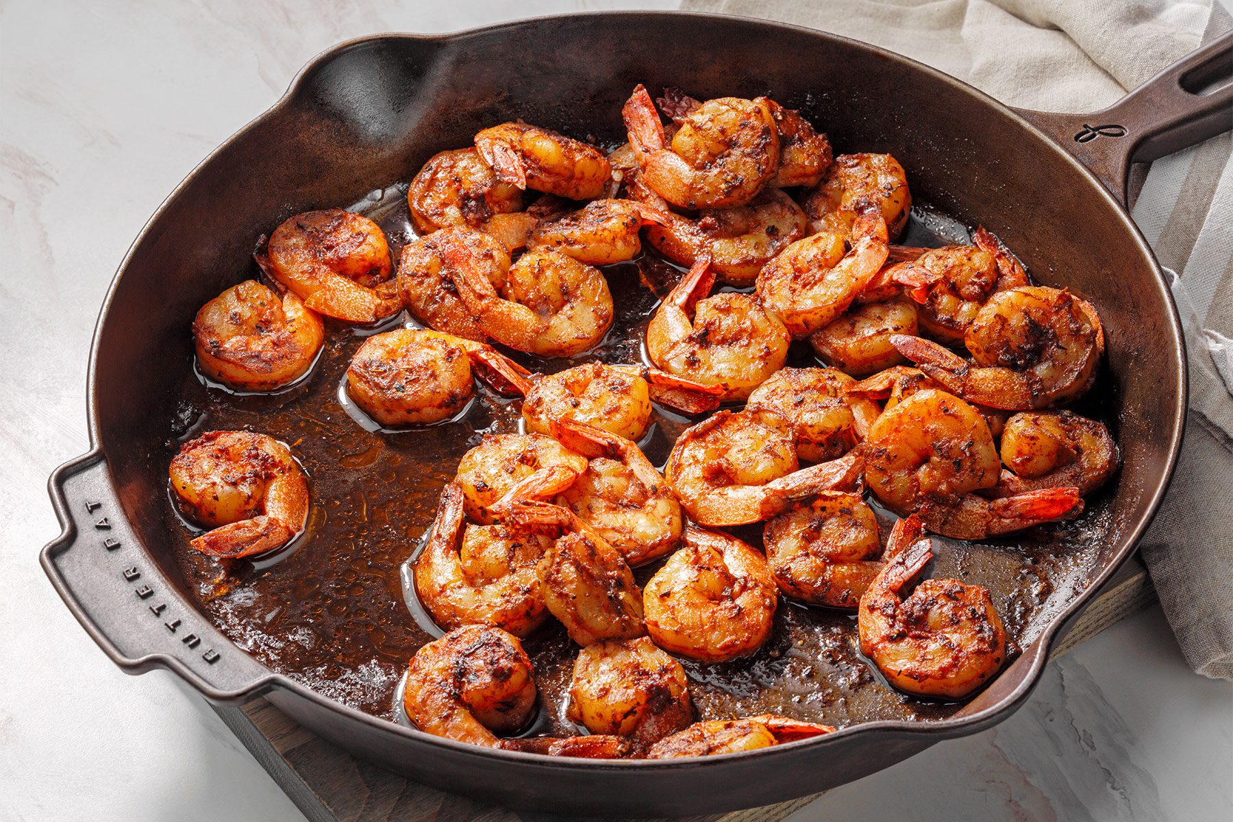 High angle shot of a large cast iron skillet; melt butter over medium heat; added shrimp; cook and stir until shrimp turn pink and edges are browned; wooden base; kitchen towel; white marble background;