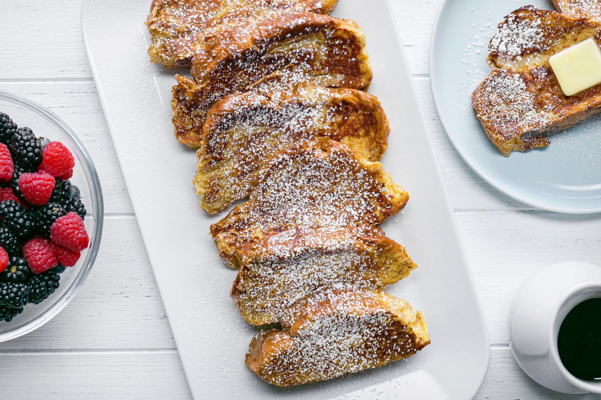 overhead shot; white background; Challah French Toast; served on white rectangular plate; Challah French Toast; served on white plate; berries in bowl; maple syrup; in beaker;