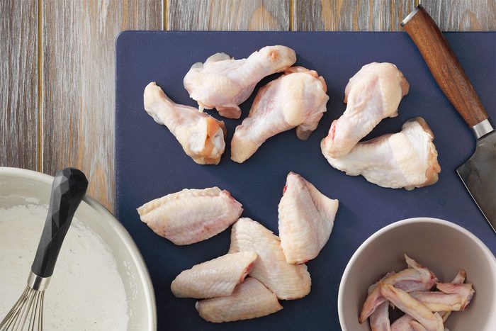 Raw chicken drumsticks and wings are shown on a dark cutting board placed on a wooden surface. Some pieces are also in a bowl and a few are next to a white pan. The ingredients are ready for preparation.