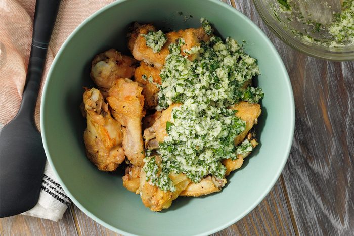 A green bowl filled with crispy chicken wings topped with a generous amount of chimichurri sauce made from parsley, garlic, vinegar, and oil, garnished with finely chopped herbs. The chicken appears golden brown, adding a contrasting color to the vibrant green sauce.