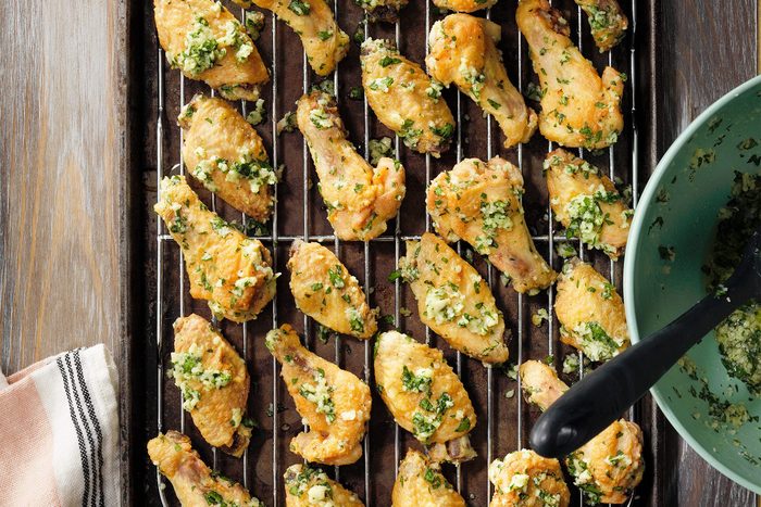 A baking sheet with a wire rack holds numerous golden brown chicken wings garnished with a generous amount of finely chopped green herbs and minced garlic, giving the wings a fresh and savory appearance. The scene suggests a freshly baked, flavorful dish.