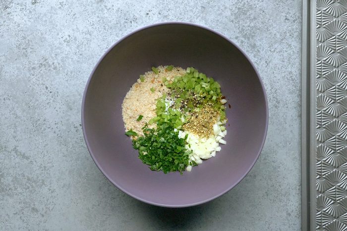 A purple bowl containing chopped green onions, celery, parsley, breadcrumbs, and various seasonings sits on a gray countertop.