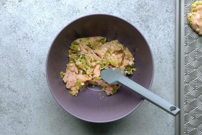 A gray bowl containing a mixture of raw chicken and green vegetables sits on a textured light gray countertop.