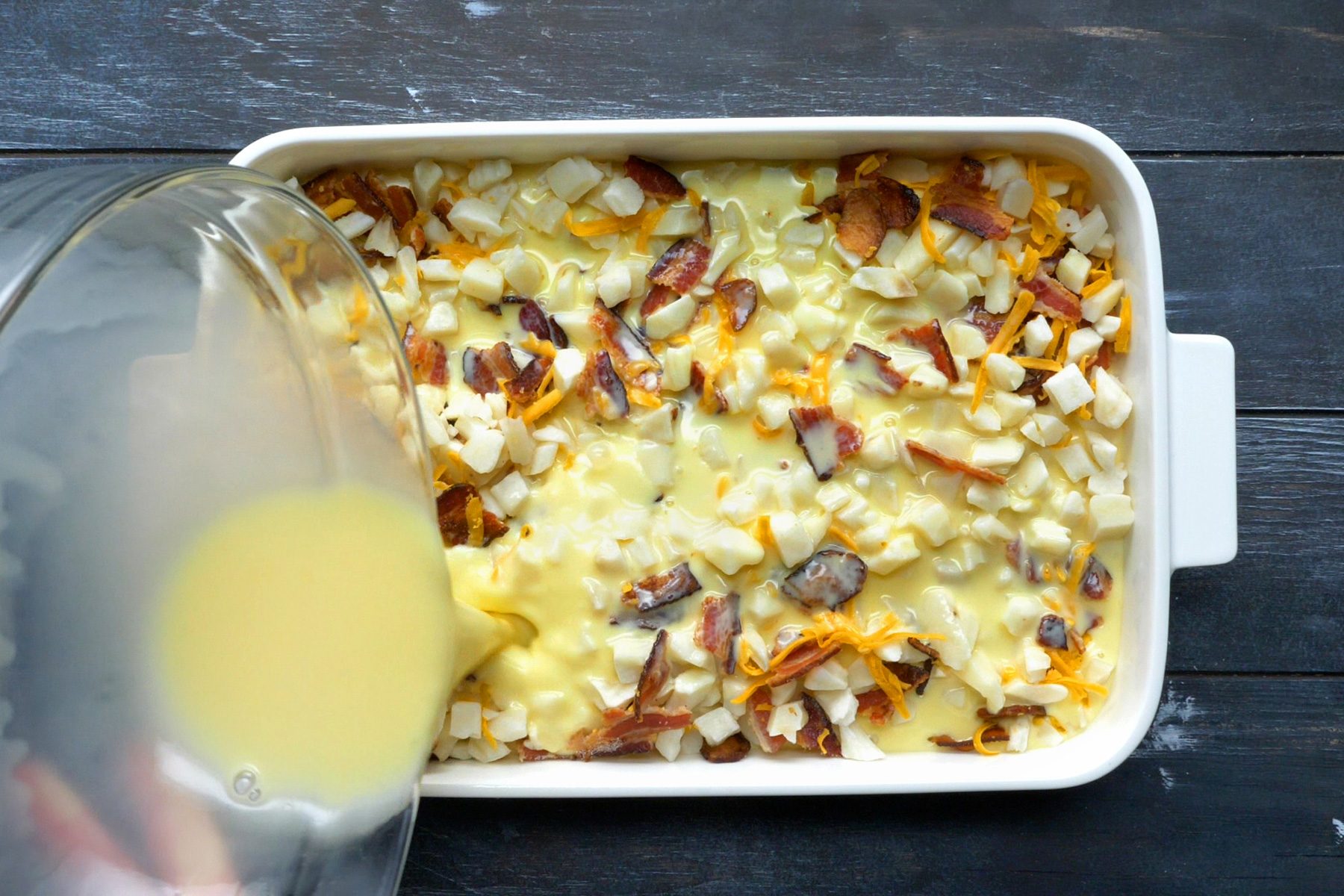 A pan of potato and bacon mixture being poured over with a wet mixture