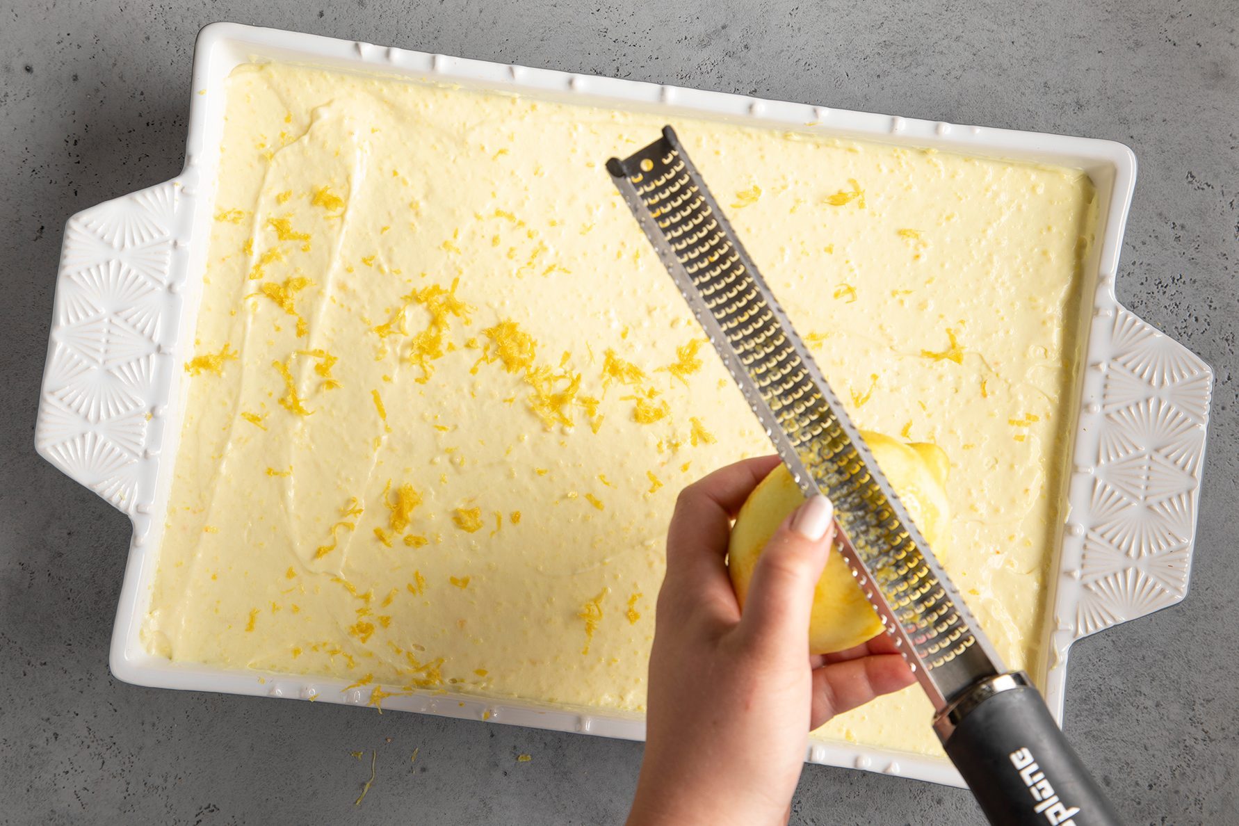 A person's hand grates lemon zest over a rectangular, baking dish filled with a creamy yellow batter.