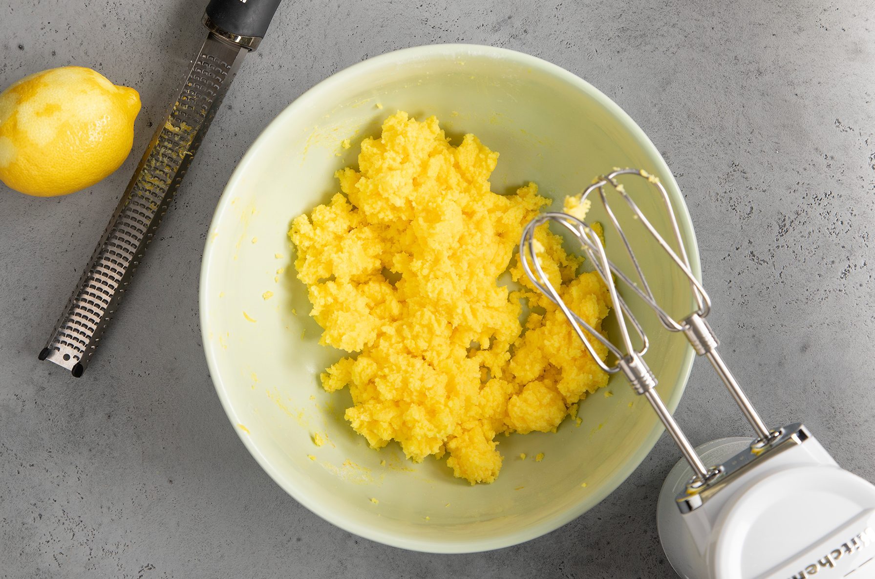 A bowl of yellow batter rests on a gray countertop. To the left, a whole lemon and a zester are placed nearby.