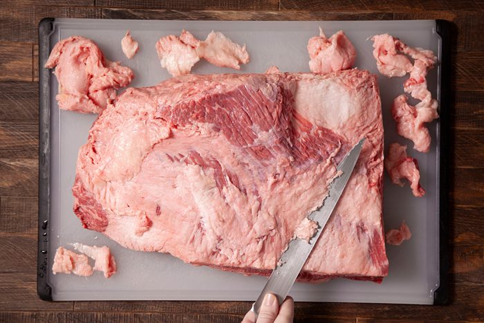 A person trimming the silverskin and fat from the top of the brisket on a cutting board.