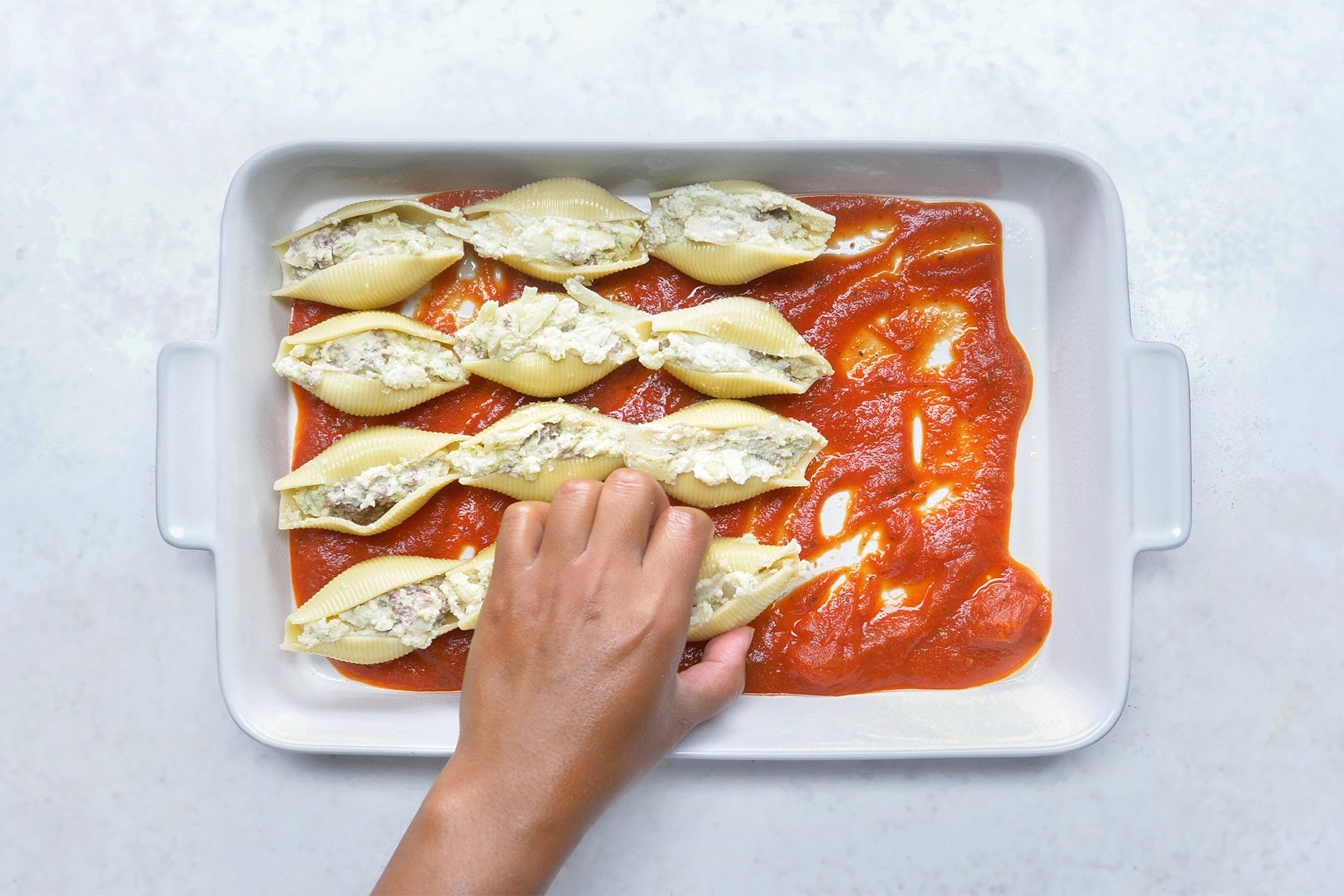 overhead shot; white background; placing filled pasta shells in baking dish; spaghetti sauce spreaded into a greased baking dish;