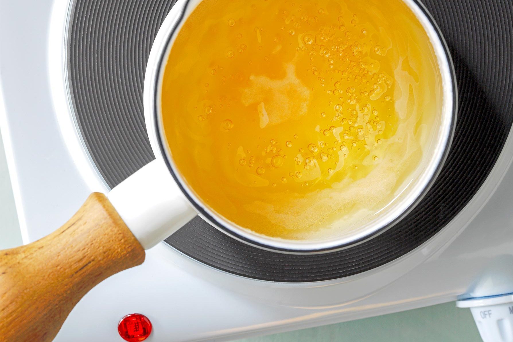 A saucepan with a white handle, filled with a light yellow liquid, sits on an electric burner with a red indicator light turned on.