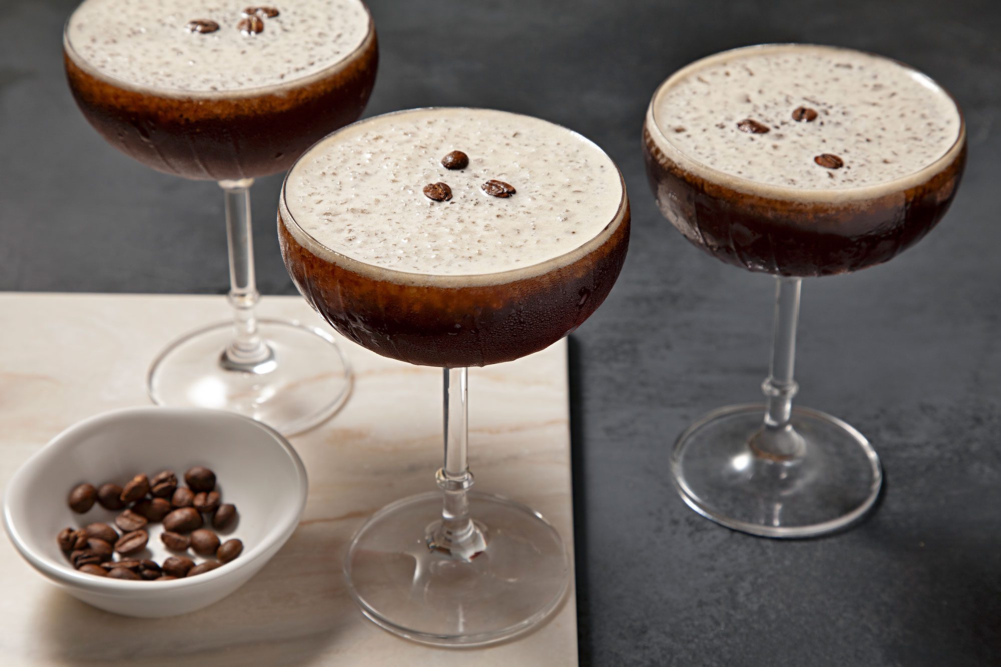 High angle view shot of Frozen Espresso Martini; in martini glasses; garnish with coffee beans; serve on wooden tray; coffee beans bowl; dark grey background;